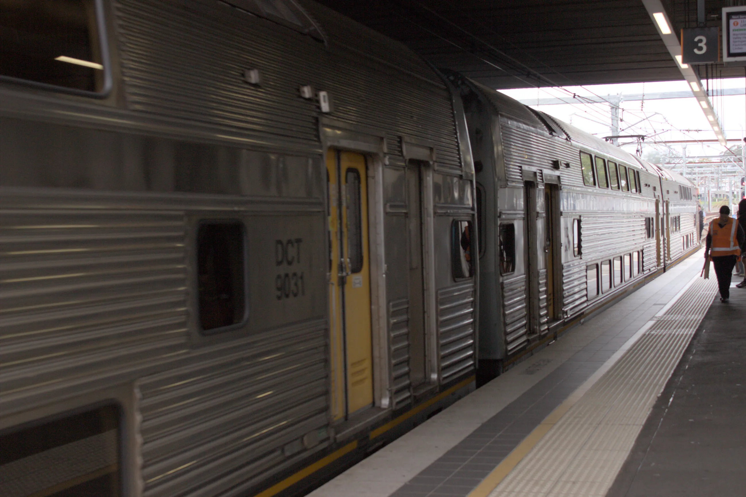 Three silver double-decker train carriages with yellow doors pulling up to a platform, with the closest carriage being identified as "DCT 9031"