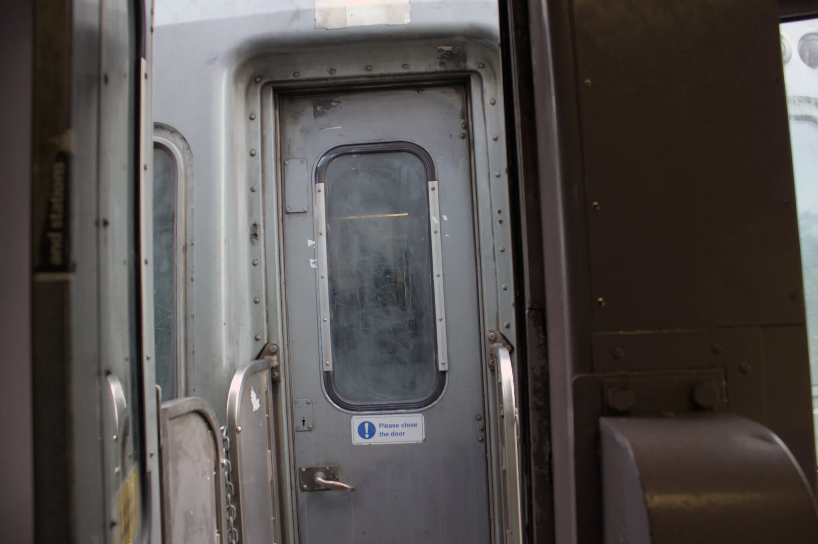 The view of the door to the other carriage over an open gangway. The door is  a greyish colour with a heavily scratched and unclear window and small handle. The gangway has shiny protective blockers on the side attached to each carriage to prevent people from falling from the train. The interior of the carriage the shot is taken from is painted purple. The lighting is mostly even, and warmer within the carriage.
