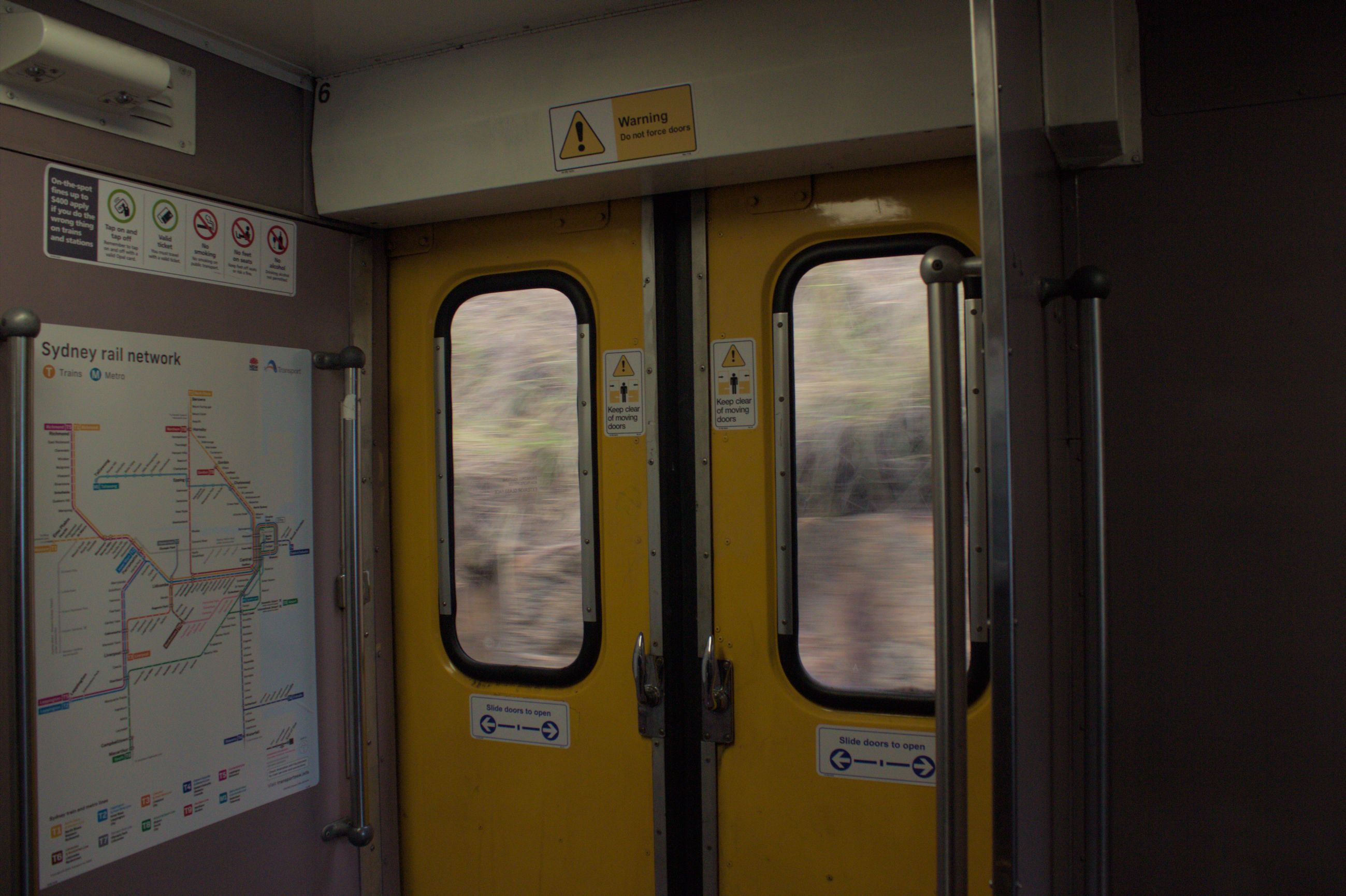 The doorway of a train carriage taken as the train is moving at speed as indicated by the blurred outside visible through the windows on the yellow doors. To the left wall next the doors a detailed map of the "Sydney rail network" is visible along with some general public transport notice stickers. There are grab handles on both sides of the doorway, and the walls are painted a unique purple colour.