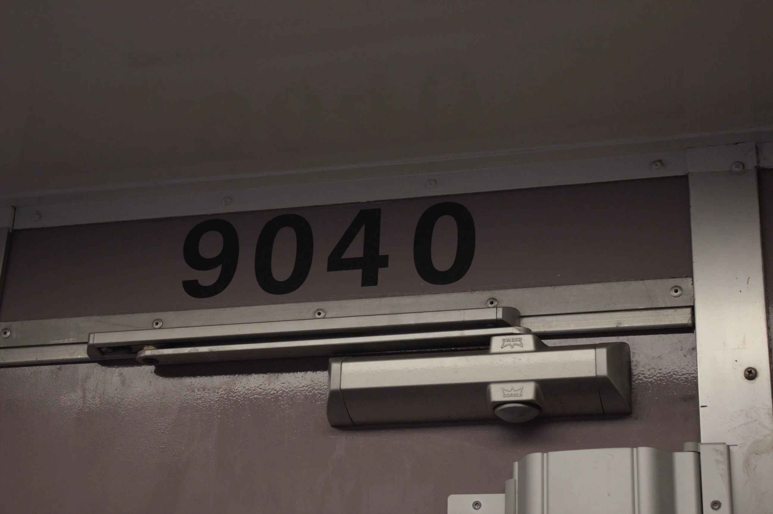 The numbers "9040" printed in large black text above the top of a doorway on  a purple wall. Only the very top of the door is visible with an automatic  door closer and replacement metal hinge.