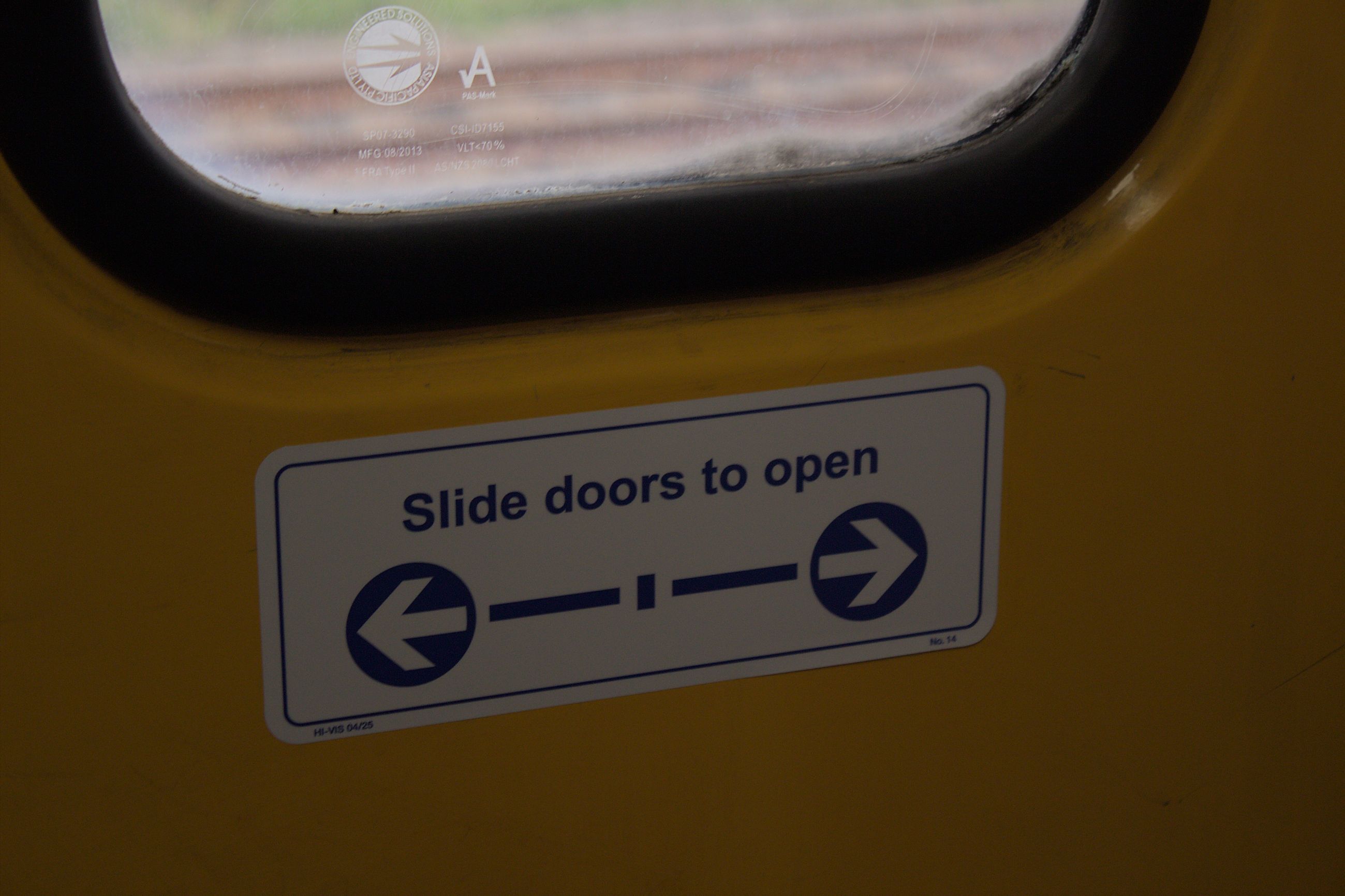 A close up of a white sticker with blue text reading "Slide doors to open" on a yellow door below a window. A related graphic with arrows and lines is below the text on the sticker. The sticker is in good condition.