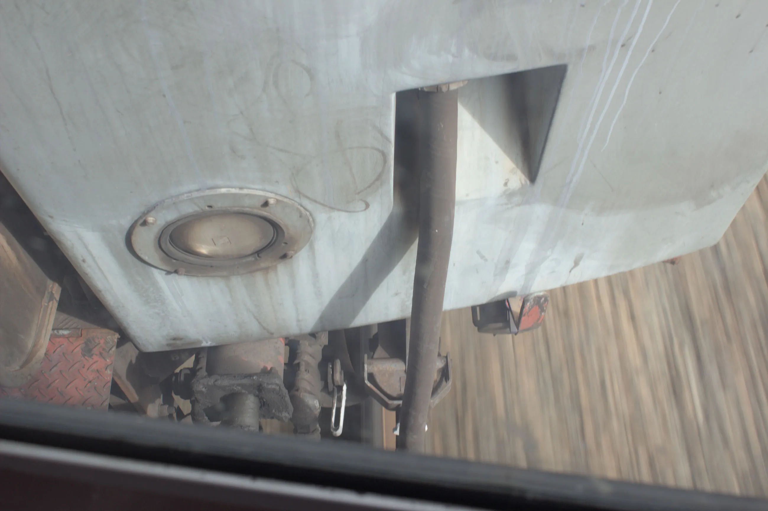 Looking out of a window down onto the gap between two carriages, with the ground and tracks visible in a blur showing that the train is moving at speed. Some  coupling equipment and a tube is visible in the frame as well, and the other  carriage is painted in a dirty grey colour. 