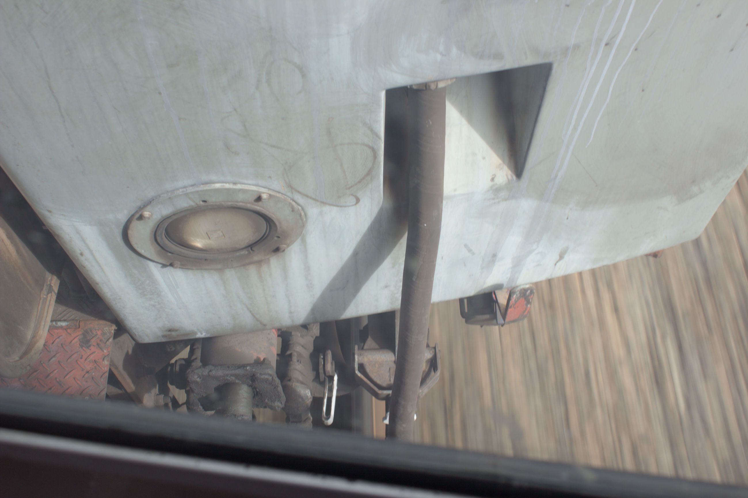 Looking out of a window down onto the gap between two carriages, with the ground and tracks visible in a blur showing that the train is moving at speed. Some coupling equipment and a tube is visible in the frame as well, and the other carriage is painted in a dirty grey colour.