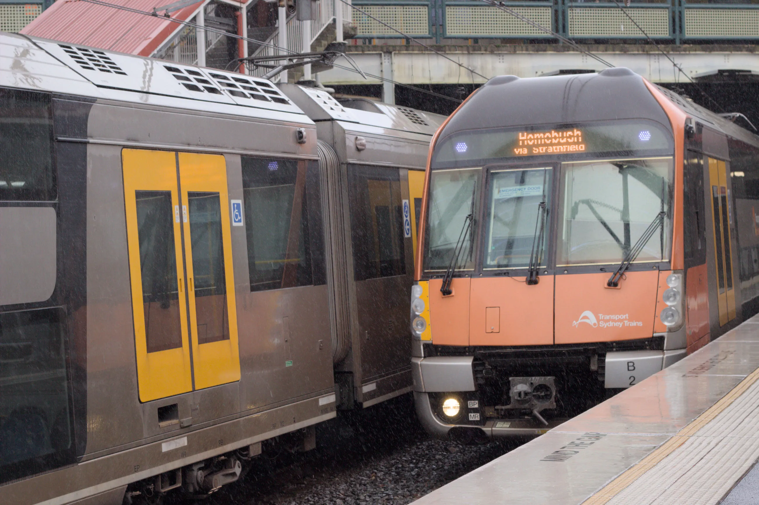 A Waratah Series 2 train with an orange and black front and a mismatched left headlight pulls into Redfern station in the rain while another B set sits on the platform opposite. The destination board reads "Homebush via Strathfield".