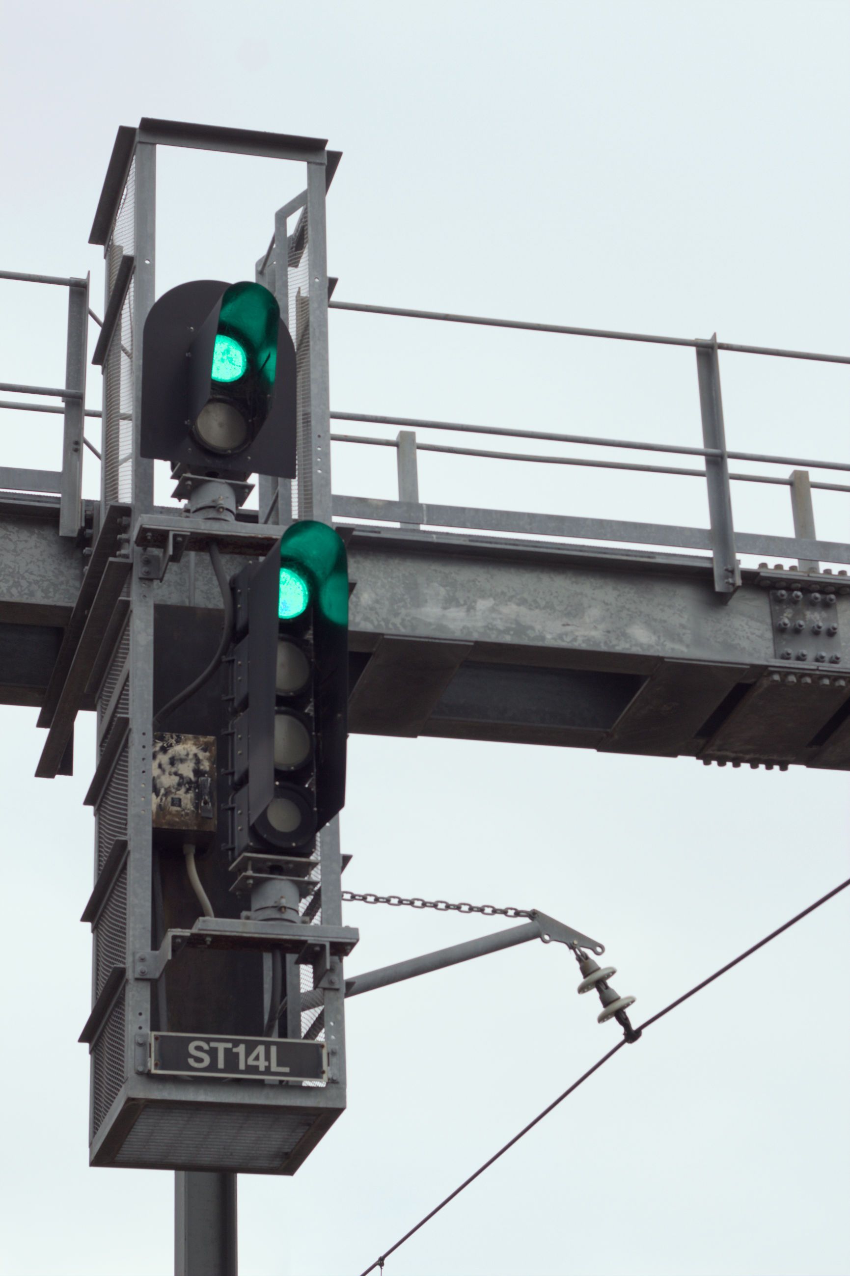 A set of railway signals mounted on an overhead steel support, made up of two sections, the top section with two lights displaying green and the bottom section with four lights also displaying green, forming a green on green "proceed" status.