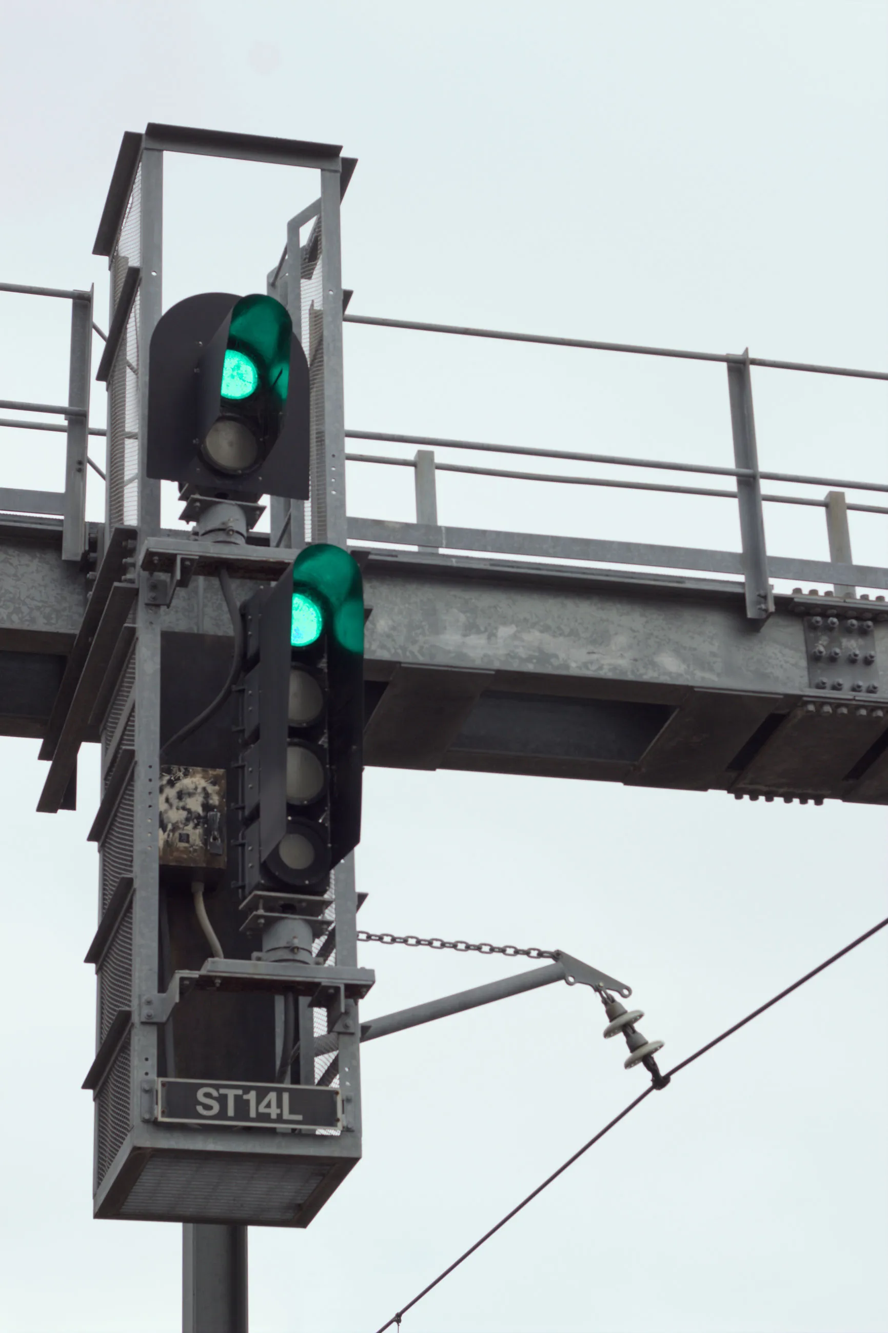 A set of railway signals mounted on an overhead steel support, made up of two sections, the top section with two lights displaying green and the bottom  section with four lights also displaying green, forming a green on green  "proceed" status.