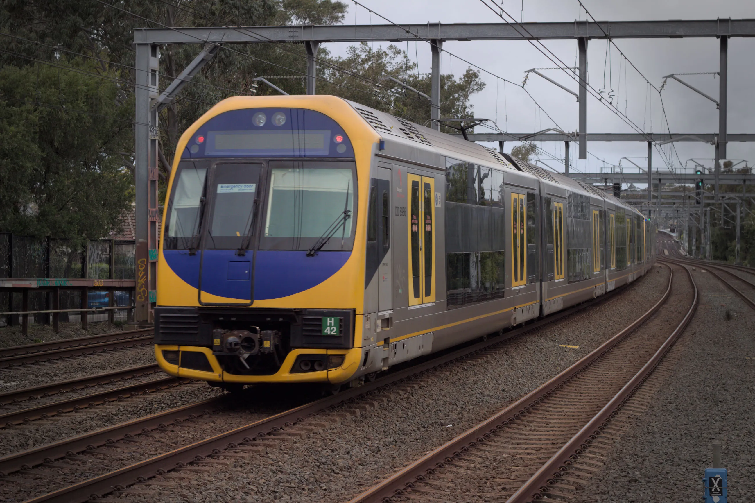 The yellow and blue rear of a double-decker silver OSCAR train moving away from the camera, passing under a wire gantry on the third from the left track out of six train tracks. The sky is overcast.