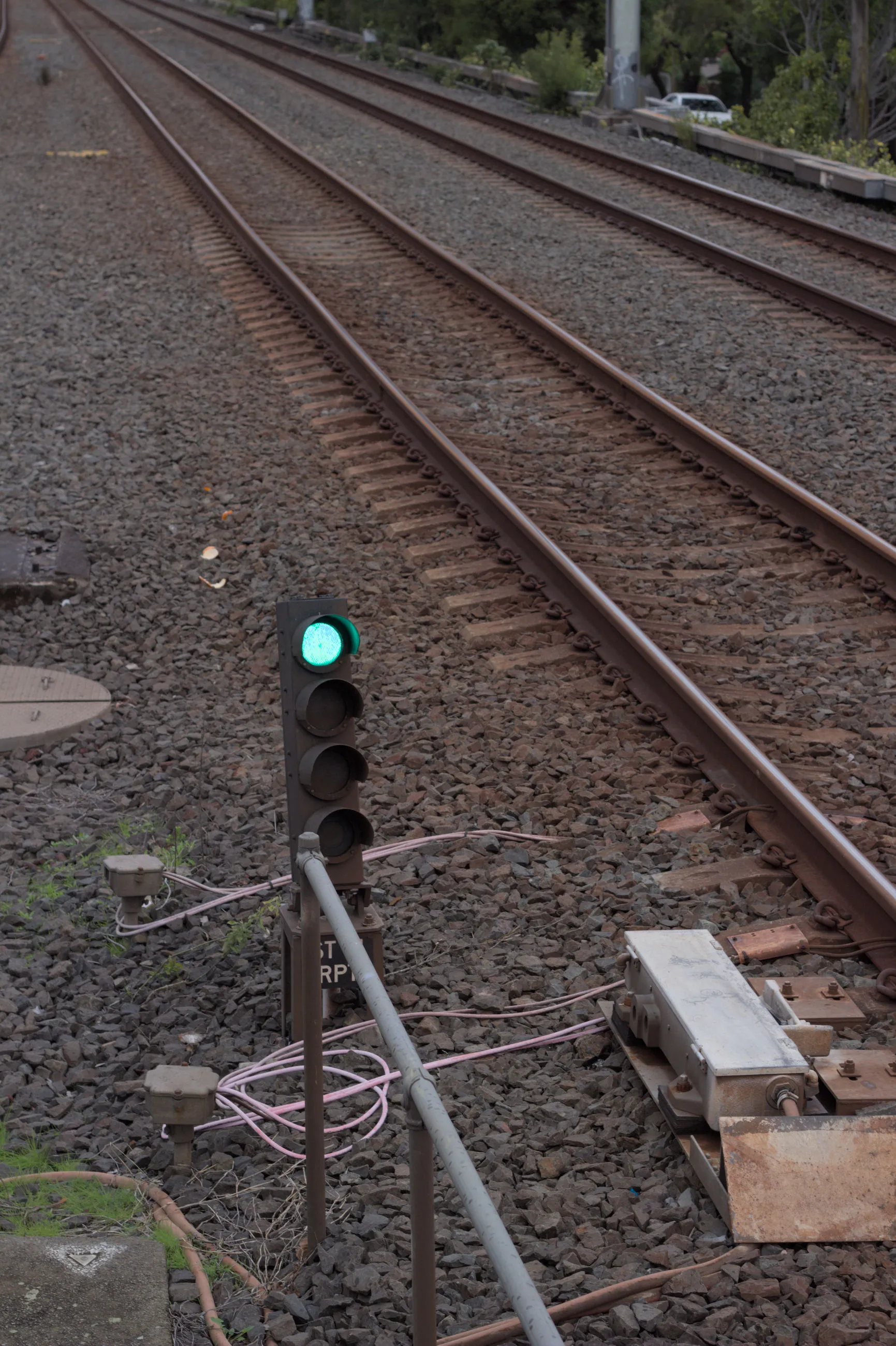 Trackside four-light railway signal with the top light illuminated green and an empty pair of tracks visible to the right of it. The white train stop directly  adjacent the signal is in the down position. 