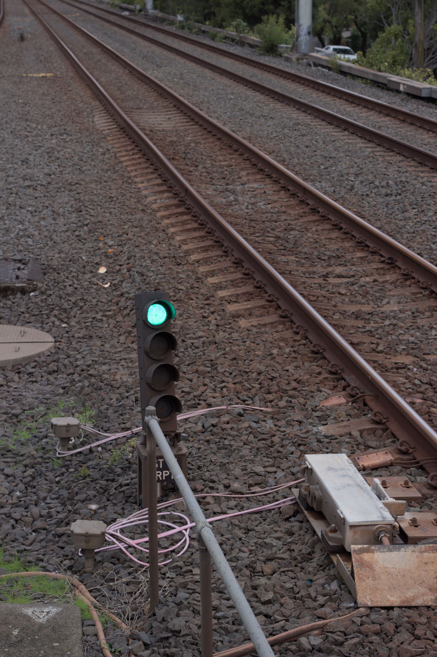 Trackside four-light railway signal with the top light illuminated green and an empty pair of tracks visible to the right of it. The white train stop directly adjacent the signal is in the down position.