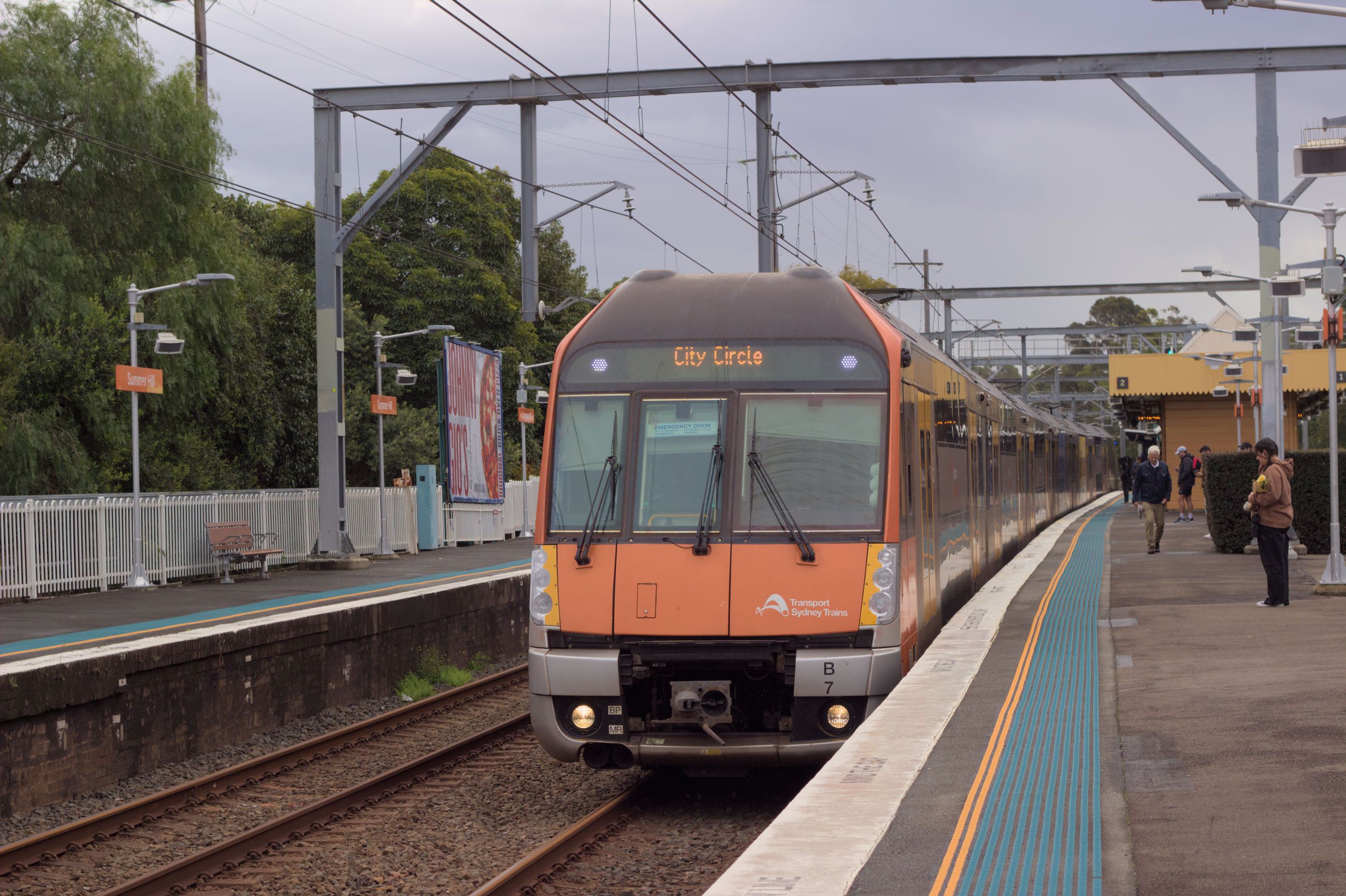 The orange and black front of a Waratah Series 2 train pulling into platform 2 at Summer Hill station. The destination board reads "City Circle". On the platform, a couple people are visible, waiting for the train to stop. The sky above is overcast.