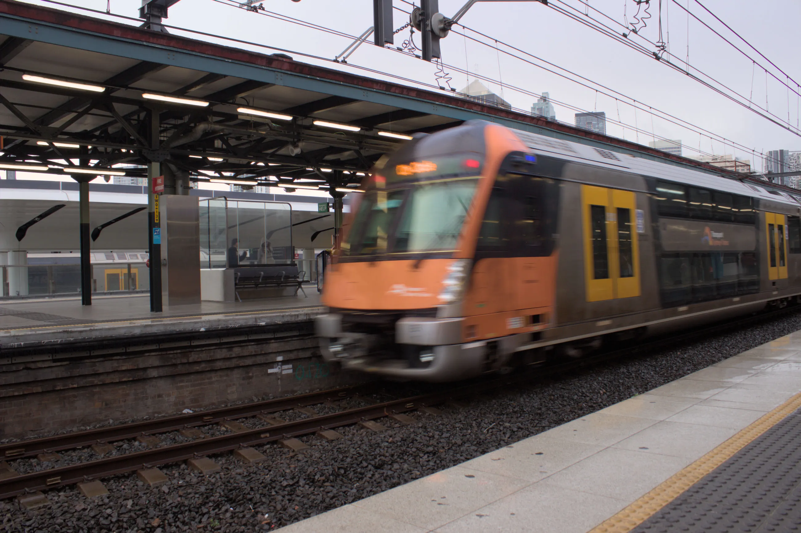 The rear of a double-decker orange and black B set with a silver body moving at speed, departing the platform opposite the camera. 