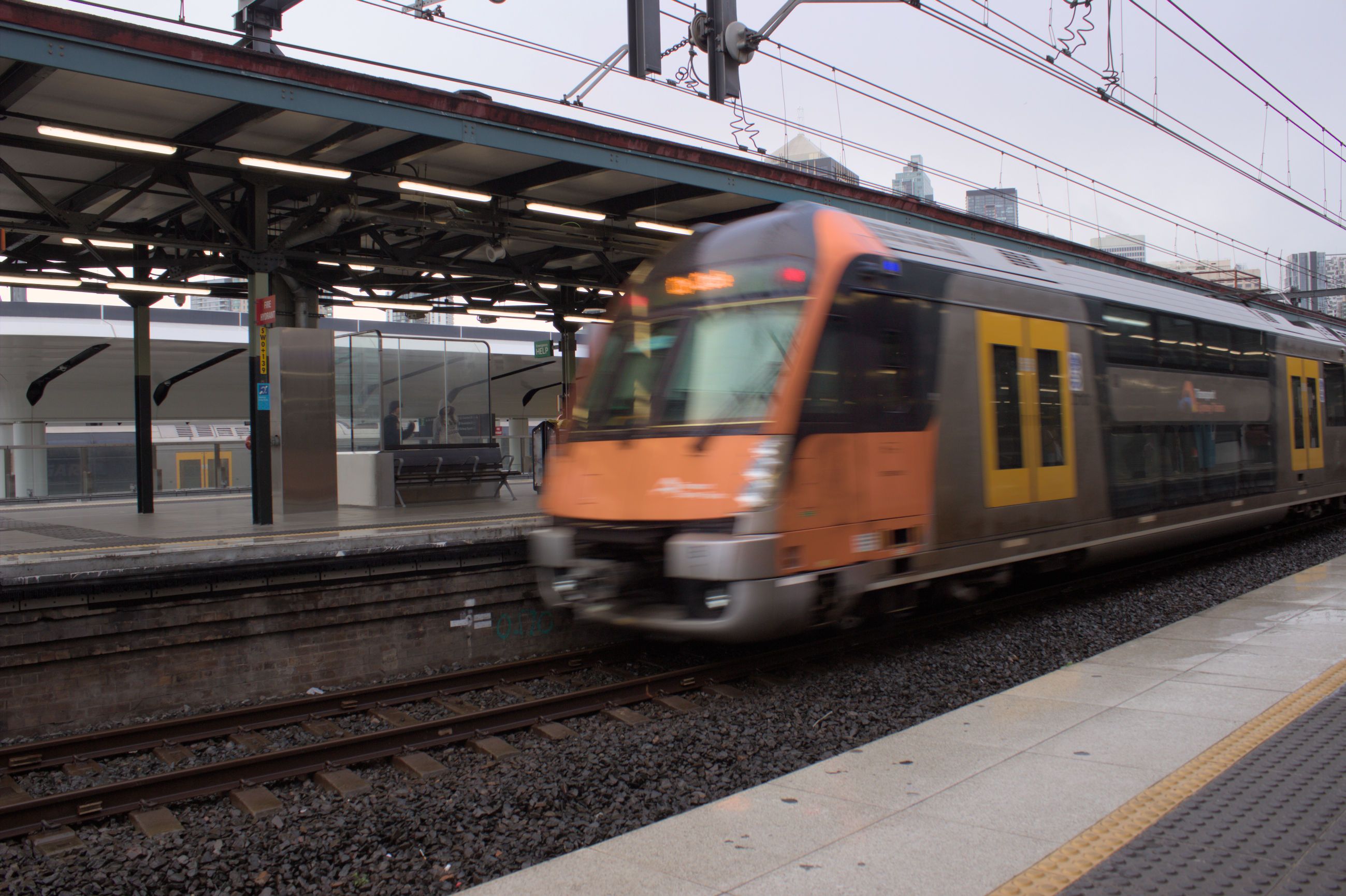 The rear of a double-decker orange and black B set with a silver body moving at speed, departing the platform opposite the camera.