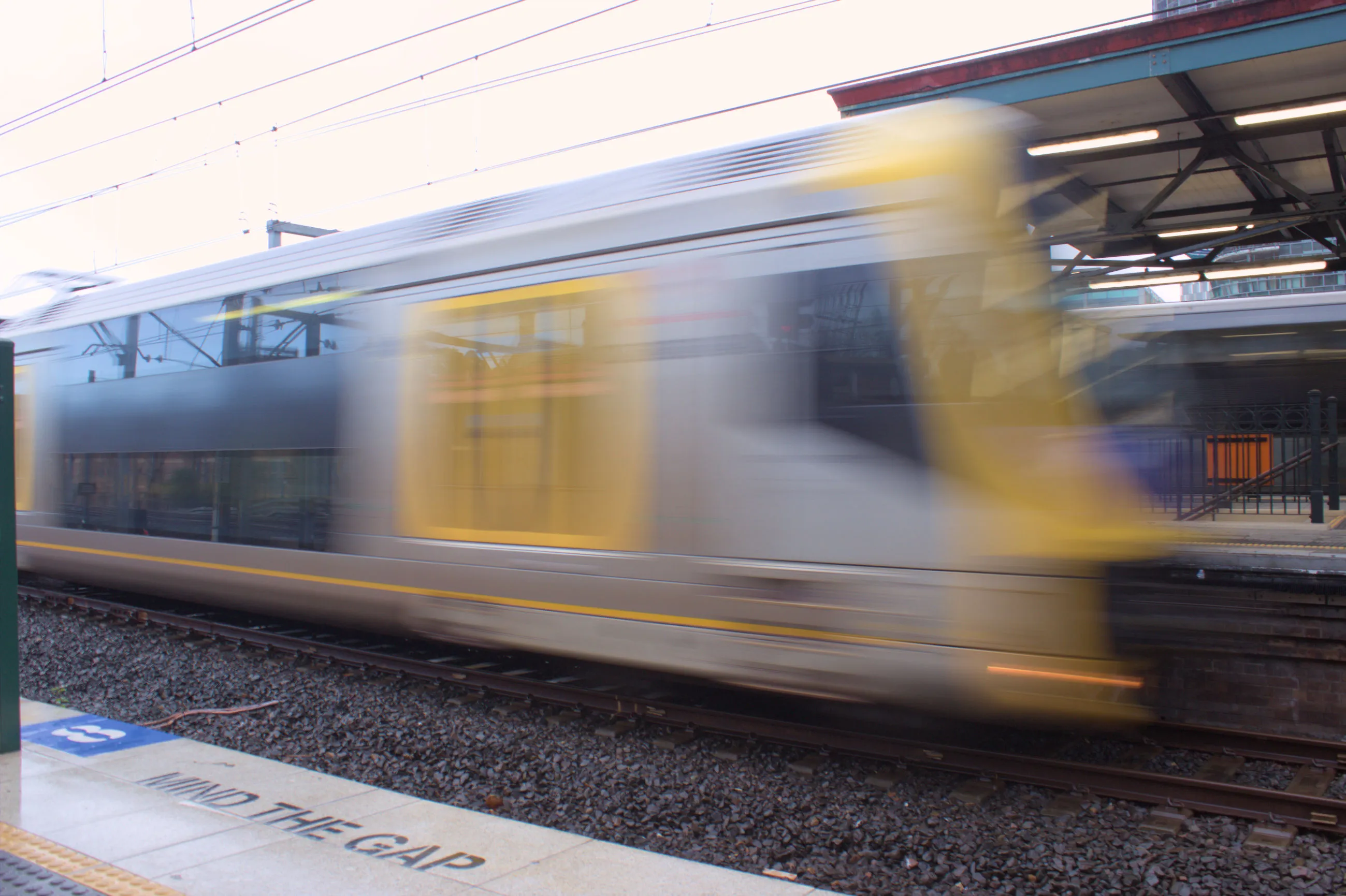 An OSCAR H set departing from the platform opposite the camera, with the train blurry from moving at speed. The train has yellow doors and the rear is a blurred yellow. The body of the train is silver.