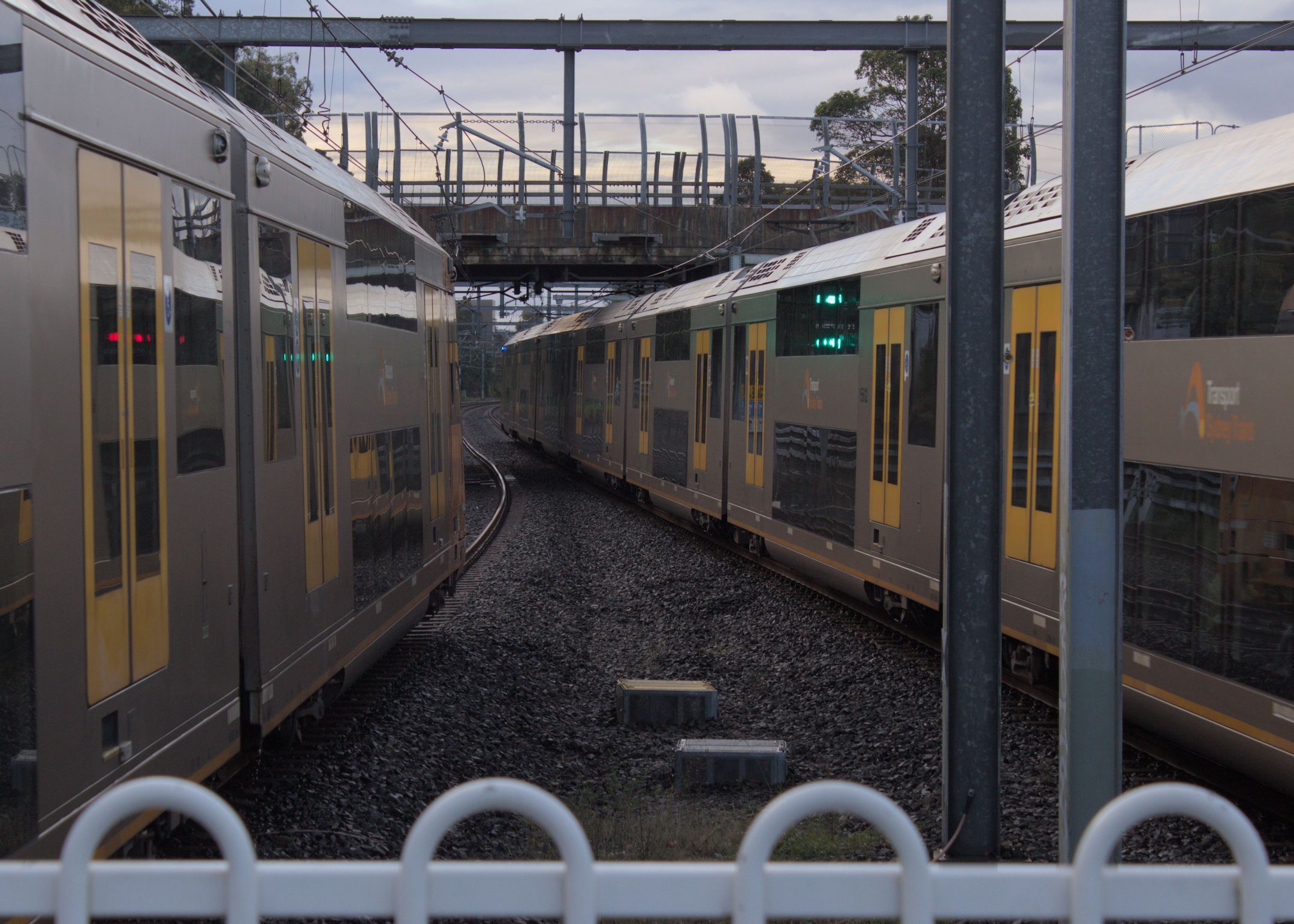Two Waratah Series 1 trains moving in opposite directions alongside each other as viewed out the end of a platform. The shot is taken as the sun is setting and the lighting is a bit dim.