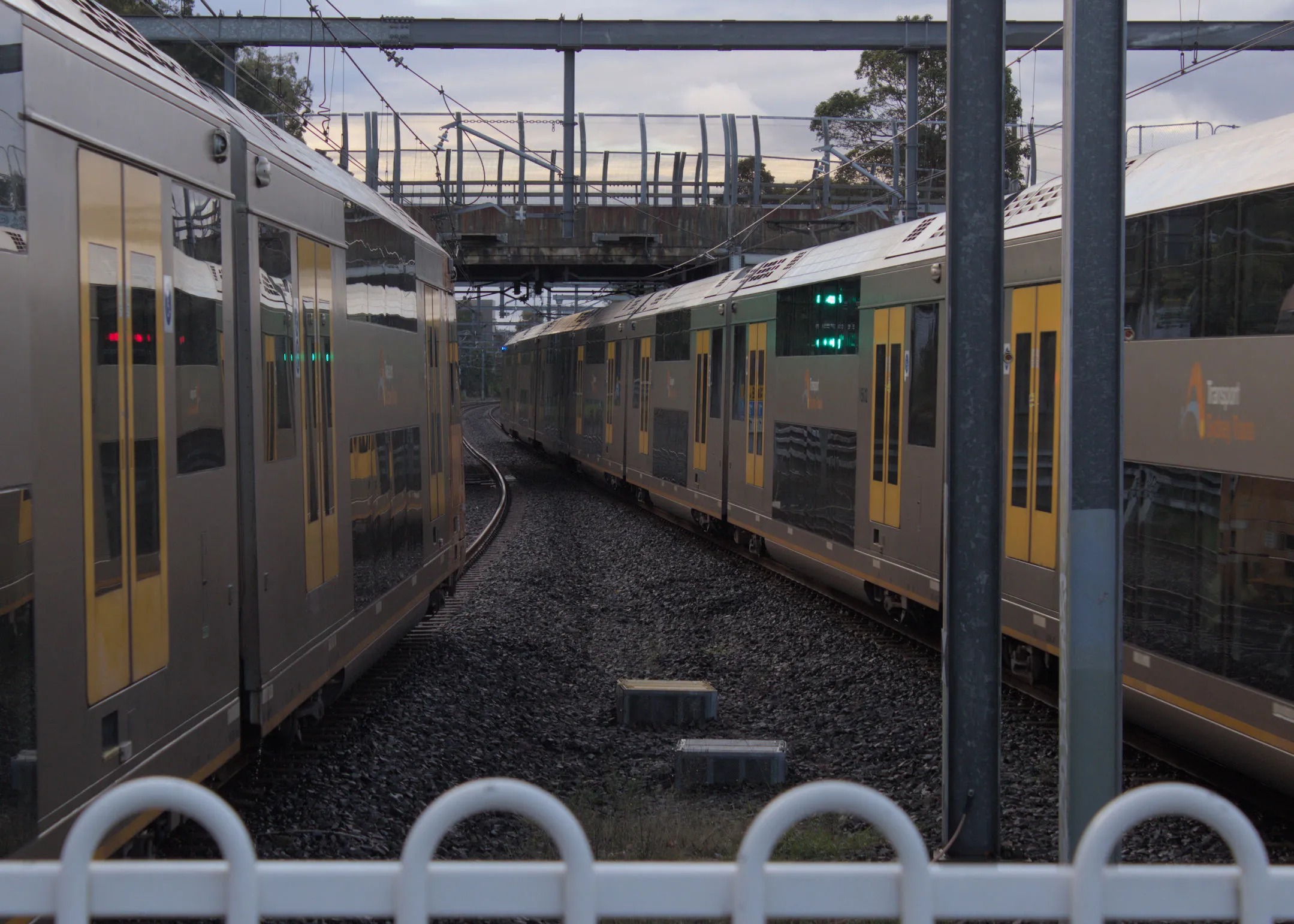 Two Waratah Series 1 trains moving in opposite directions alongside each other as viewed out the end of a platform. The shot is taken as the sun is setting and the lighting is a bit dim.