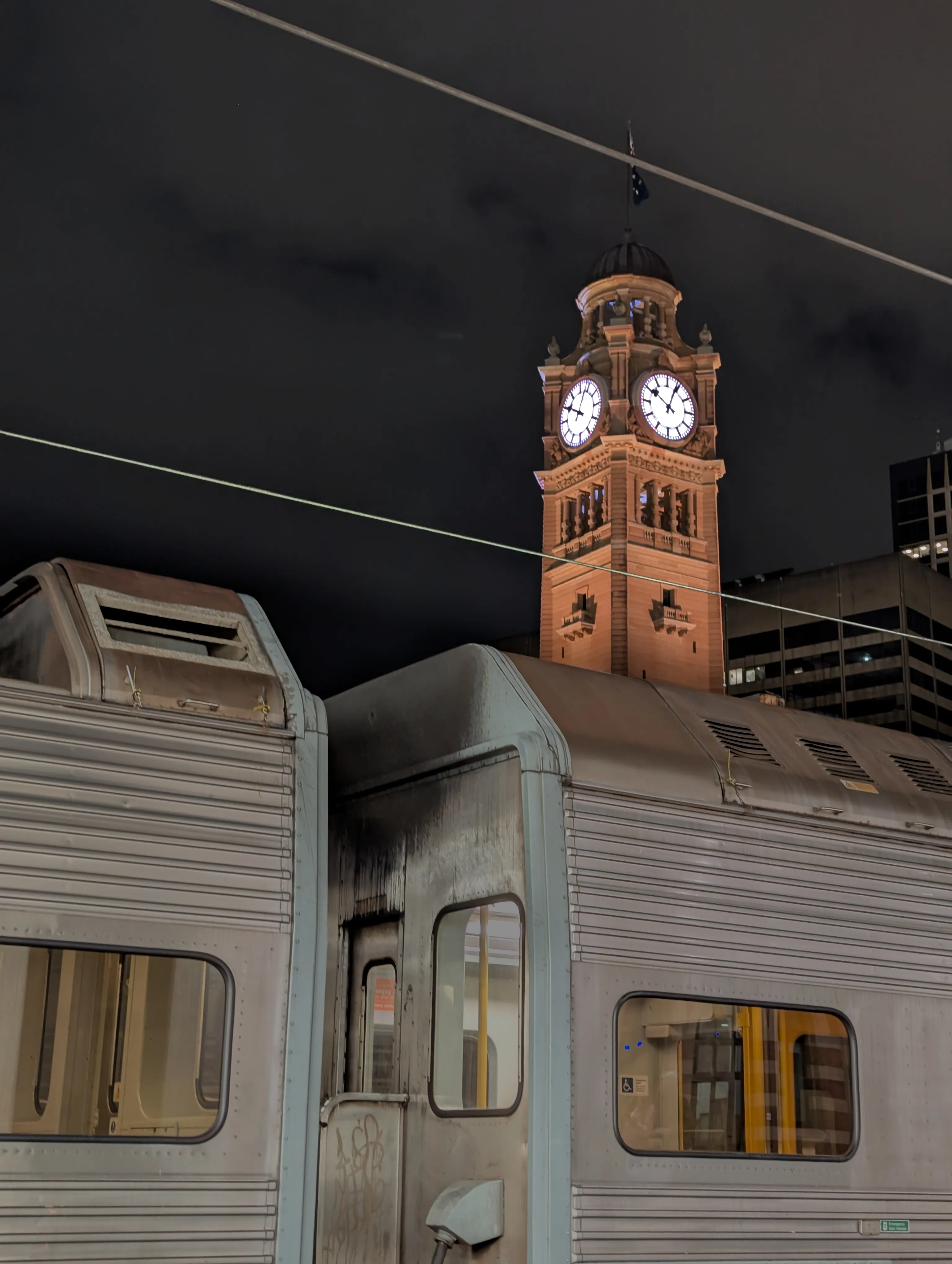 A silver suburban K set train stopped at Central at night, with the bright sandstone coloured clock tower of the station prominently visible in the background. The clock shows 10:04
